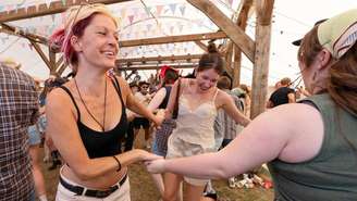 Um grupo de pessoas dá as mãos e dança em roda dentro de uma grande tenda decorada com bandeirolas coloridas, em um ambiente de festival. Em primeiro plano, três mulheres sorriem e parecem aproveitar a música, enquanto outras pessoas conversam e assistem a uma apresentação ao fundo.