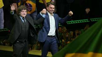 Javier Milei e Flávio Bolsonaro de mãos dadas para o alto. 