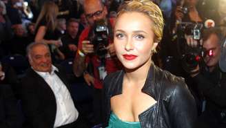 Hayden Panettiere, rodeada de fotógrafos, usa o cabelo preso, batom vermelho e um vestido verde com uma jaqueta de couro preta por cima.