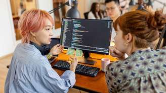 A foto ilustrativa mostra uma profissional de tecnologia com cabelo rosa sentada em uma mesa, gesticulando enquanto conversa com um colega que apoia o queixo na mão. Outros funcionários aparecem ao fundo, em um ambiente de escritório, e a linguagem de programação está visível na tela do computador.