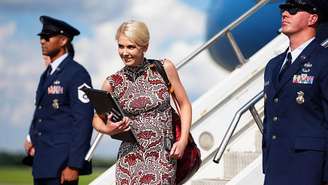 Natalie Harp desce do Air Force One com um vestido estampado sem mangas, sorrindo com um laptop nas mãos