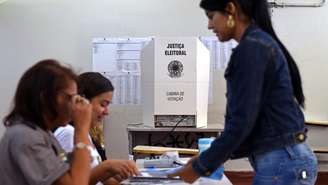 Duas mesárias atendem eleitora em local de votação com a cabina de votação ao fundo
