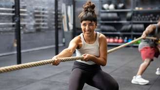 Uma jovem de leggings pretas e blusa sem mangas, com os cabelos escuros presos, puxa com força uma corda de cabo de guerra em uma academia com piso cinza. Ao fundo, veem-se prateleiras de pesos e um homem (desfocado) de short vermelho segurando a mesma corda