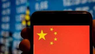 Mão segura um telefone celular com a bandeira da República Popular da China na tela, tendo ao fundo uma tela grande com números desfocados