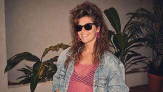 Um retrato ao estilo dos anos 80, gerado por inteligência artificial, mostra uma jovem sorridente com óculos escuros, mechas loiras, um longo permanente castanho com volume na raiz, jaqueta jeans azul-escura, blusa rosa de tela, brincos de argola e batom, em frente a um cenário de vasos de plantas.