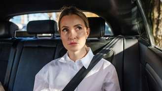 Uma mulher vestindo uma camisa branca está sentada no banco de trás de um carro. Ela está olhando pela janela e usando cinto de segurança. Ela não parece feliz.