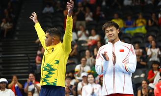 Wendell Belarmino, de agasalho amarelo e detalhes verde, ergue os braços e comemora no pódio. Ao seu lado, está um chinês, com agasalho branco, que aplaude 