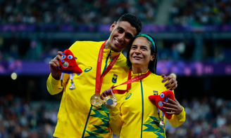 Gabriel Garcia e Jerusa Geber estão com a medalha de ouro no peito e seguram a Phryge, a mascota dos Jogos. Ele a abraça, vestindo uniforme amarelo, com detalhes em verde 