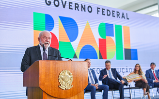 Em primeiro plano, Lula discursa em púlpito; ao fundo, ministros estão sentados em cadeiras e há painel com logo do governo federal.