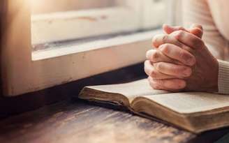 Pessoa com as mãos fechadas sobre uma bíblia próxima a janela
