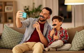 Homem ao lado do filho, sentados em um sofá, ambos com um bigode de papel, tirando uma foto