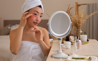 Mulher coreana fazendo seu skincare, com fita rosa nas olheiras, com toalha na cabeça e no corpo, na frente de um espelho redondo de mesa