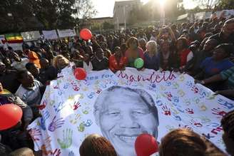 Pessoas abrem um cartaz com a imagem de Nelson Mandela durante as comemorações pelo seu 95º aniversário, em frente à clínica onde o ex-presidente da África do Sul está internado, em Pretória. A saúde de Mandela melhora a cada dia e ele agora é capaz de sentar-se por alguns minutos, informou sua filha mais nova à emissora estatal SABC. 18/07/2013.