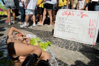 De topless e com uma maça na boca, uma ativista ficou deitada como se fosse o ingrediente principal de um prato de comida; cartaz exibe o que seria o "menu da Fifa"