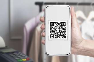 Mão segura um smartphone com um QR code na tela. Ao fundo e desfocado, há uma máquina de cartão de crédito à esquerda e uma arara com camisas penduradasmelhor jogo para jogar no pixbetcabides à direita.