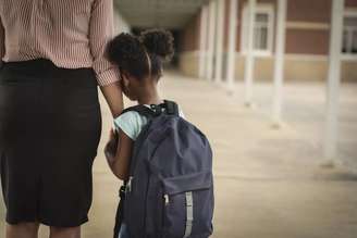 Imagem mostra uma menina negra e pequena, com uma mochila nas costas, abraçada ao braço de uma mulher adulta.