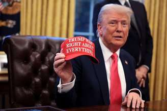 Trump no Salão Oval da Casa Branca segurando boné vermelho com a estampa "Trump estava certo sobre tudo" em inglês