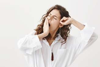 Studio portrait of tired or exhausted young businesswoman yawning and stretching while covering mouth with hand, standing with closed eyes over gray background. Girl have not got enough sleep.