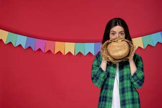 Mulher segurando chapéu de palha em frente ao rosto, atrás dela bandeirinhas de festa junina coloridas.