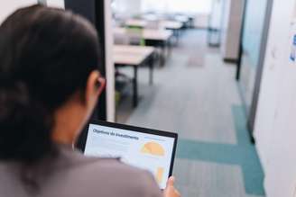 Mulher, de óculos e cabelos pretos, aparece de costas trabalhando em um tablet