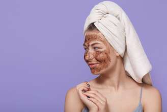 Portrait of female model applies chocolate mask on face, has positive expression, looks aside, wears towel on head after bath, poses over lilac wall with, uses beauty product for skin care.