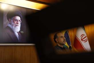 No detalhe, o embaixador do Irã no Brasil, Abdollah Nekounam, durante conversa com jornalistas, sob a imagem do aiatolá Ali Khamenei, morto em bombardeio americano e israelense