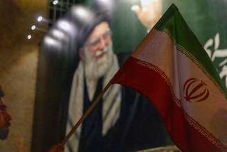 Uma bandeira nacional iraniana é hasteada em frente a uma faixa com a imagem de Ali Khamenei durante uma vigília em memória do líder supremo do Irã, Ali Khamenei, após a mídia estatal iraniana confirmar sua morte em 1º de março de 2026, em Teerã, Irã.