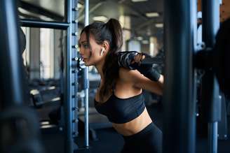 Side crop of brunette female bodybuilder using wireless headphones and doing squats in multipower smith machine. Srtong woman with muscular body training legs in gym. Concept of bodybuilding.
