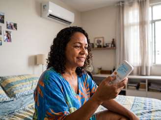 Imagem mostra uma mulher negra de cabelos cacheados sentada na cama com um controle do ar-condicionado na mão.