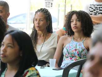 Imagem mostra duas mulheres negras observando atentamente um interlocutor enquanto há outras mulheres em volta.