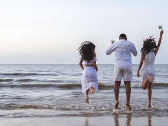 Imagem mostra duas mulheres e um homem à beira da praia vestindo branco.