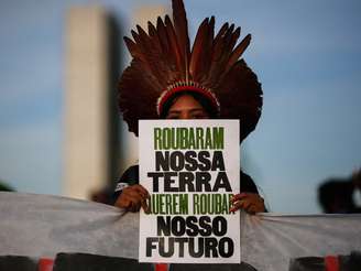 A imagem mostra uma mulher indígena segurando uma placa escrita "roubaram nossa terra, querem roubar nosso futuro", em manifestação contra o marco temporal.