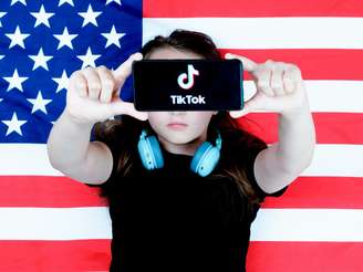 Imagem centraliza uma criança tampando os olhos com um celular na vertical, que mostra a logo do TikTok no centro da tela. No fundo da foto, uma bandeira dos Estados Unidos.