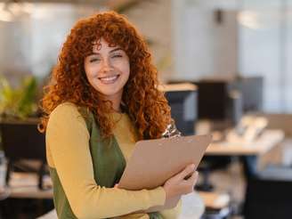 Imagem mostra mulher ruiva da Geração Z sorrindo em ume escritório com uma prancheta na mão. Ela possui cabelos enrolados e usa camisa de manda longa amarela e um colete verde por cima.