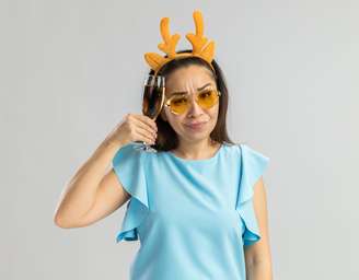 young woman in blue top wearing funny rim with deer horns and yellow glasses holding glass of champagne looking at camera with skeptic smile on face standing over white background
