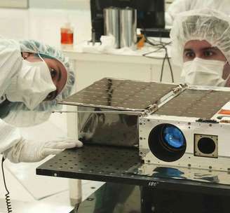 Dois cientistas usando máscaras, luvas, coberturas para a cabeça e trajes limpos brancos trabalham em um instrumento em um laboratório.