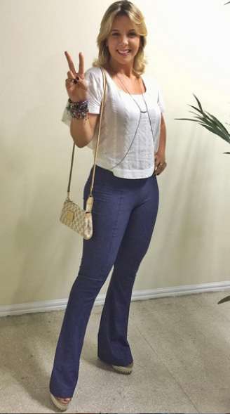 Carla Perez usa calça azul, tipo flare, com blusa branca curta. A calça marca mais do que devia e o comprimento da blusa acaba valorizando demais os quadris. A bolsa pequena e o colar, conhecido como body chain, estão fora de contexto no look